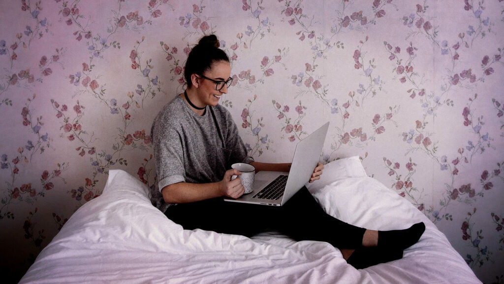 Image of Woman sitting on her bed reading from laptop with a cup of coffee.